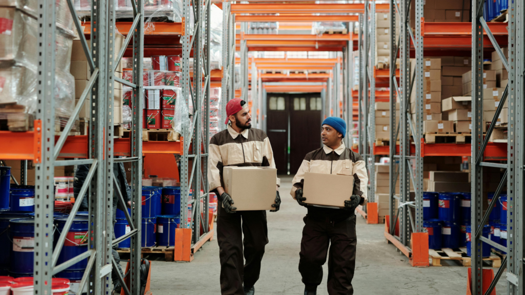 Warehouse employees carrying inventory boxes through a commercial storage aisle with tall industrial shelving, illustrating warehouse operations, logistics management, and commercial facility security in a Bay Area distribution center.